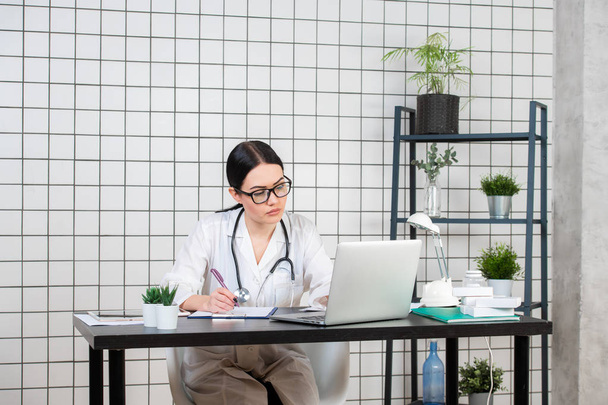 Hermosa mujer médico escribiendo mientras usa el ordenador portátil en el escritorio en la clínica
 - Foto, Imagen