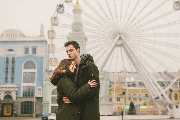 Theme love and holiday Valentines Day. pair of Caucasian heterosexual lovers in winter together gloomy weather embrace against background of Ferris wheel in town square. The guy gently hugs the girl. - Photo, Image