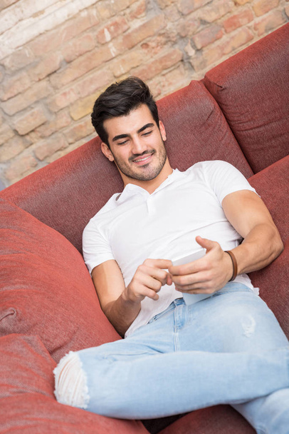 A smiling young man laying on a sofa and using his smartphone - Фото, изображение