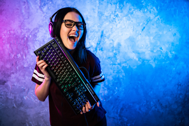 Bella amichevole Pro Gamer Streamer ragazza in posa con una tastiera nelle sue mani, indossando occhiali. Attraente ragazza geek con colori retrò neon cool in background. - Foto, immagini