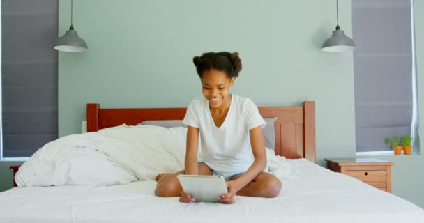 Front view of black girl using digital tablet in bedroom at home. Black girl sitting on bed in bedroom 4k - Séquence, vidéo