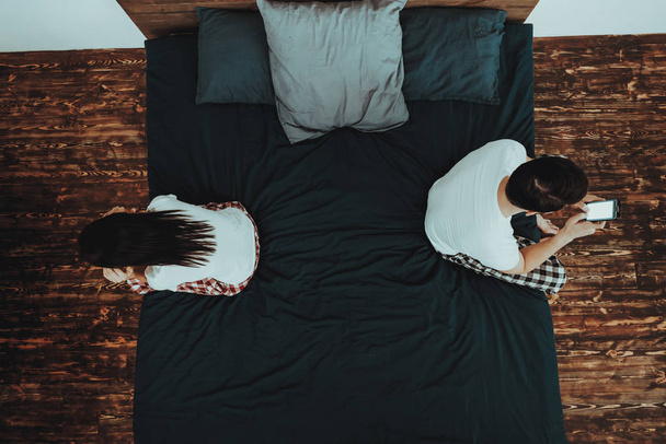 Couple is Sitting on Bed Back to Back. Couple is Young Beautiful Woman and Man. Guy is Using Cellphone. Girl is Covering Her Ears. People is Wearing Pajama Pants and T-Shirts. Home Interior. Top View. - Foto, Imagem
