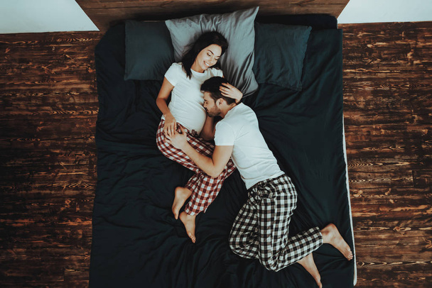 Couple is Lying on Bed. Couple is Young Beautiful Woman and Man. Girl is Pregnant. Man is Listening Belly of Woman. Persons is Happy and Smiling. People in Home Interior. Top View. - Фото, изображение