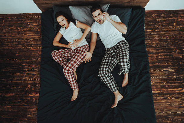 Couple is Lying on Bed. Couple is Young Beautiful Woman and Man. Woman is Gives Birth and Touching Her Belly. Anxious Man is Calling on Cellphone to Hospital. People in Home Interior. Top View. - Photo, image