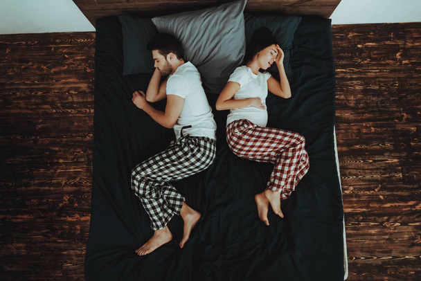 Couple is Lying and Sleeping on Bed. Couple is Young Beautiful Woman and Man. Woman is Pregnant and Feeling Headache. People is Wearing Pajama Pants and T-Shirts. People in Home Interior. Top View. - Foto, afbeelding