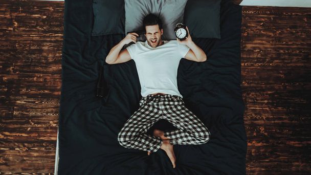 Man is Lying on Bed and Screaming. Young Beautiful Caucasian Man. Person is Wearing Pajama Pant and T-Shirt. Man is Holding Pistol at the Head and Alarm Clock. Person in Home Interior. Top View. - Foto, immagini