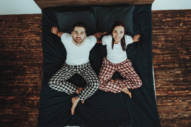 Couple is Sitting on Bed. Couple is Young Beautiful Woman and Man. Persons is Looking Up and Smiling. People is Wearing Pajama Pants and T-Shirts. People is Located in Home Interior. Top View. - Foto, Bild