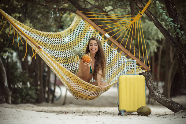 Tanned beautiful girl is sitting in hammock on the beach with yellow suitcase - Фото, изображение
