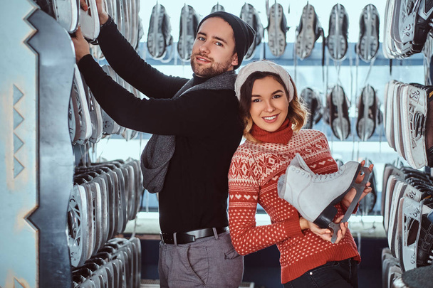 Young couple wearing warm clothes standing near rack with many pairs of skates, choosing his size - Photo, Image