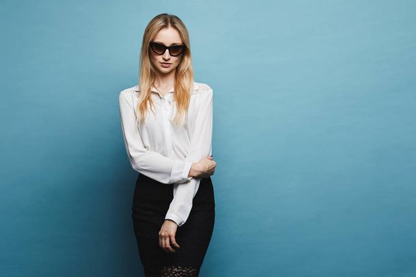 Beautiful, fashionable and sexy blonde model girl in stylish sunglasses and in a white blouse and black tight skirt posing at the blue background in a studio, isolated - Fotó, kép