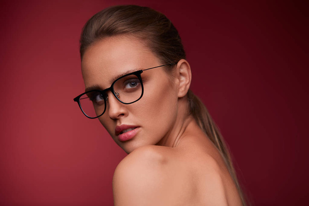 Attractive woman in square eyeglasses looking at camera on red background. Portrait of young beautiful blonde girl.Black frame spectacles.Vision correction,optical shop,glasses store,eyewear concept. - Foto, imagen