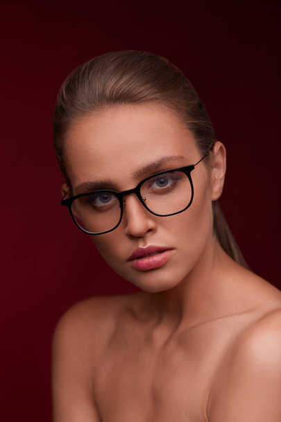 Attractive woman in square eyeglasses looking at camera on red background. Portrait of young beautiful blonde girl.Black frame spectacles.Vision correction,optical shop,glasses store,eyewear concept. - Foto, afbeelding