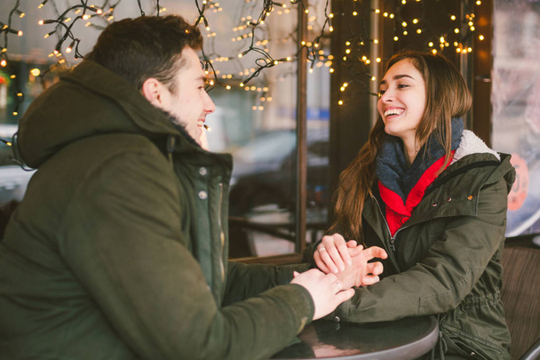 Theme love holiday Valentines Day. pair college students, Caucasian heterosexual lovers in winter, sit table of street cafe against the background window lights. Emotion romance of happiness and love. - Foto, Imagen
