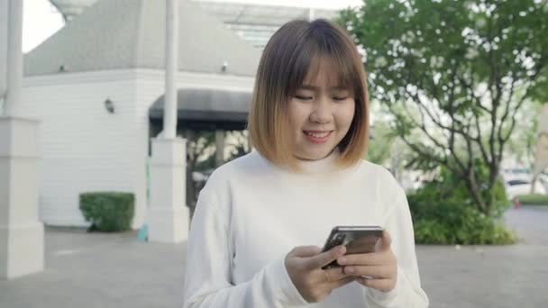 fröhliche asiatische Touristenbloggerin mit Touchscreen-Technologie am Smartphone, während sie abends auf der Straße in der Innenstadt spaziert. Lifestyle Rucksack touristisches Reisekonzept. - Filmmaterial, Video