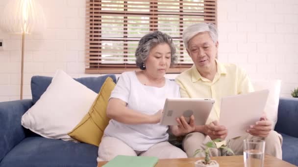 Asian elderly couple using tablet watching TV in living room at home, couple enjoy love moment while lying on sofa when relaxed at home. Enjoying time lifestyle senior family at home concept. - Footage, Video