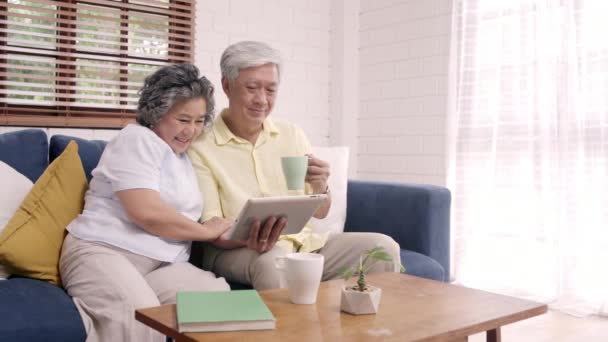 Asian elderly couple using tablet and drinking coffee in living room at home, couple enjoy love moment while lying on sofa when relaxed at home. Enjoying time lifestyle senior family at home concept. - Footage, Video