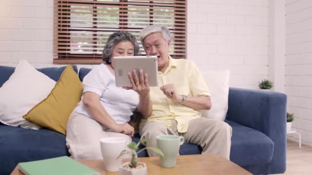Asian elderly couple using tablet watching TV in living room at home, couple enjoy love moment while lying on sofa when relaxed at home. Enjoying time lifestyle senior family at home concept. - Filmagem, Vídeo