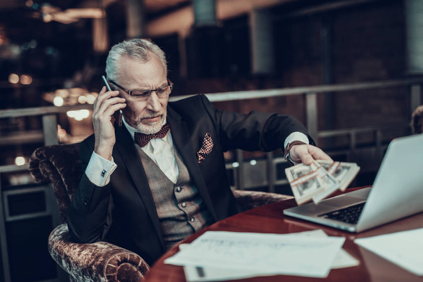 Businessman Using Laptop. Old Businessman . Man is Wearing in Black Suit. Experienced Entrepreneur. Strict Man. looking at Laptop. Working Late. talking on Phone. holding stack of money - Foto, imagen