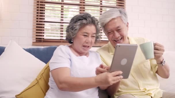 Asian elderly couple using tablet and drinking coffee in living room at home, couple enjoy love moment while lying on sofa when relaxed at home. Enjoying time lifestyle senior family at home concept. - Footage, Video
