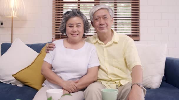 感じて幸せ笑顔としながらカメラをアジアの老夫婦は、自宅のリビング ルームでソファの上リラックスします。時間ホーム コンセプト シニア家族ライフ スタイルを楽しんでいます。カメラ目線の肖像画. - 映像、動画