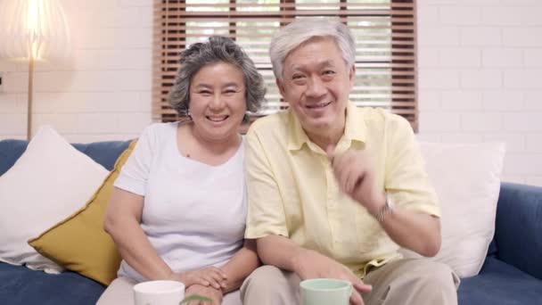 Asian elderly couple using smartphone video conference with grandchild while lying on sofa in living room at home. Enjoying time lifestyle senior family at home concept. Portrait looking at camera. - Footage, Video