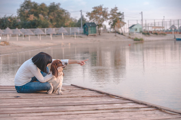 Portret z piękną, młodą kobietę, grając z psem nad rzeką. Szczęśliwa kobieta siedząca na molo z jej pies Basset Hound. Kobieta z szczeniak.  - Zdjęcie, obraz