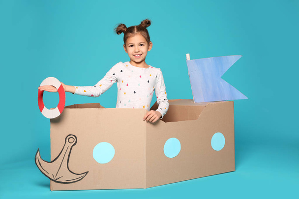 Cute little girl playing with cardboard boat on color background - Photo, Image
