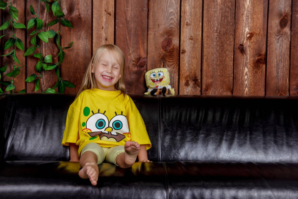 Portrait of cute European girls sitting in a yellow spongebob t-shirt on a black leather couch in the Studio with a background of wooden brown boards - Photo, Image
