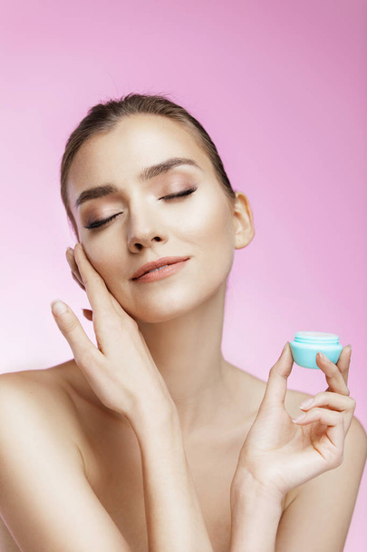 Beauty portrait of girl with perfect nude make-up. Holding cream and touching her face. Pink background.  Beauty salon. Head and shoulders, studio, indoors  - Zdjęcie, obraz