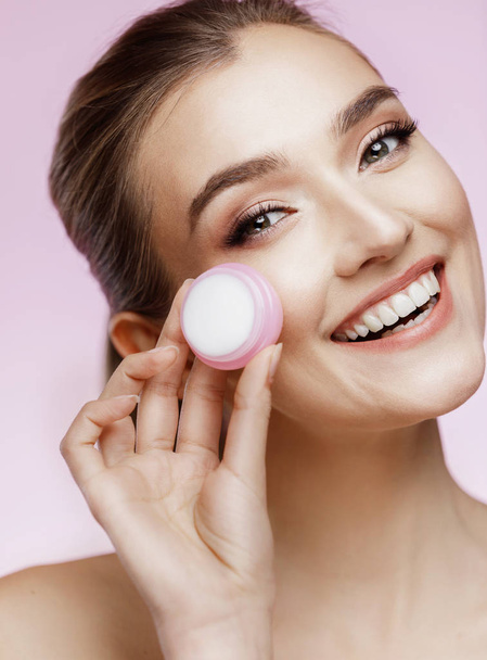 Beauty portrait of girl with perfect nude make-up. Holding cream and touching her face. Pink background.  Beauty salon. Head and shoulders, studio, indoors  - Fotografie, Obrázek