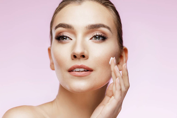 Beauty portrait of girl with perfect nude make-up. Holding cream and touching her face. Pink background.  Beauty salon. Head and shoulders, studio, indoors  - Фото, изображение