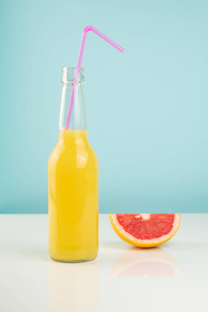 Orange drink and slice of grapefruit on white and blue background. Minimalistic image of citrus juice bottle and fruit at sparse bright environment. - Fotoğraf, Görsel
