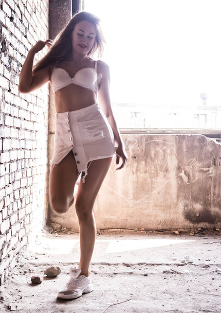 Sexy young woman in a short white skirt posing near a brick wall in sunlight. blurred light - Фото, зображення