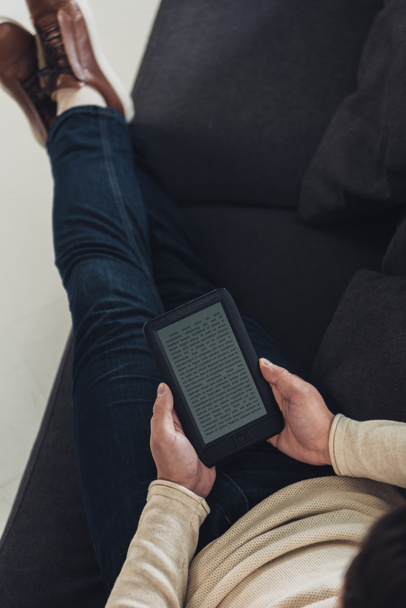 Draufsicht eines Mannes, der mit einem E-Book auf einem Sofa mit überkreuzten Beinen sitzt - Foto, Bild
