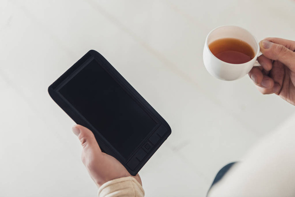 levágott nézet üres képernyő és a csésze tea, kezében az ember otthon eBook - Fotó, kép