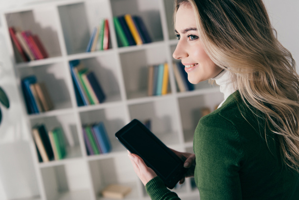 fröhliche Frau hält E-Book mit leerem Bildschirm in der Nähe des Bücherregals zu Hause  - Foto, Bild