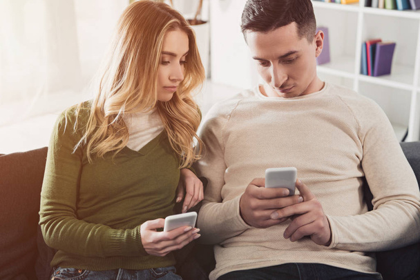 woman looking at smartphone in hands of boyfriend while sitting on sofa at home - Photo, Image