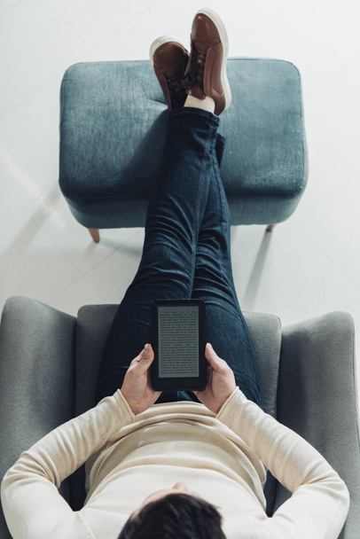 κάτοψη του άνδρα να κρατάει e-αναγνώστη, ενώ κάθεται στην πολυθρόνα  - Φωτογραφία, εικόνα