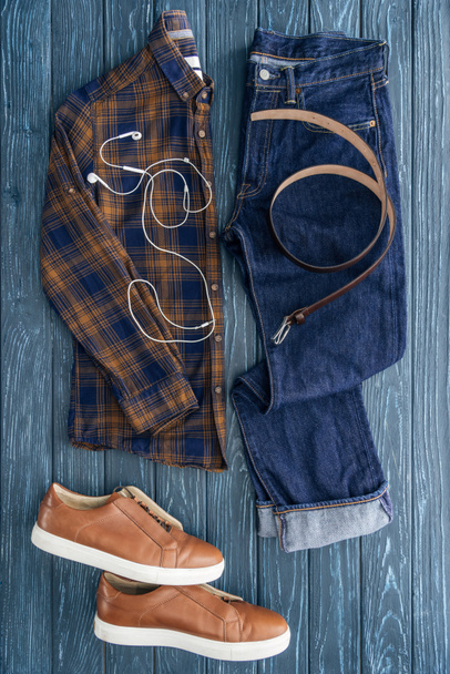Top view of jeans, checkered shirt and earphones on wooden background - Photo, Image