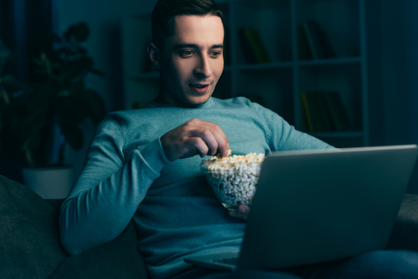 handsome man watching movie on laptop and holding bowl with popcorn at home - Fotoğraf, Görsel