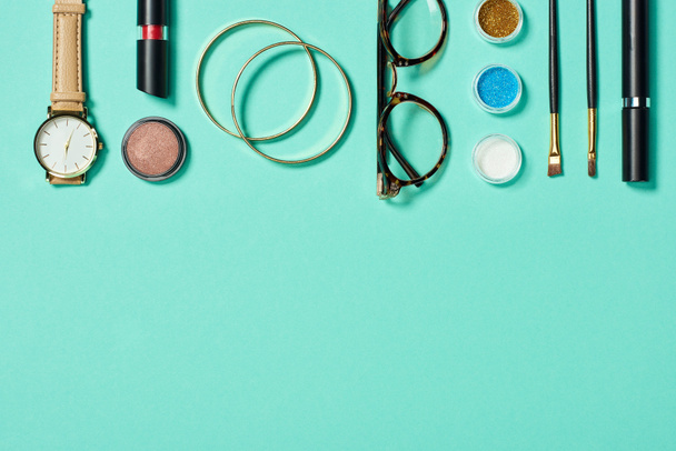 Top view of watch, lipstick, bracelets, glasses, eyeshadow, blush, cosmetic brushes and mascara on turquoise background - Photo, Image