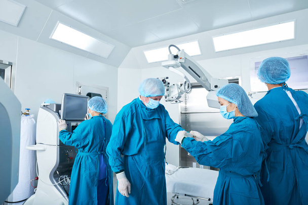 Młody asystent chirurgiczny pomoc doświadczonego chirurga, aby umieścić na Rękawiczki lateksowe przed wykonaniem zabiegu w blok operacyjny - Zdjęcie, obraz
