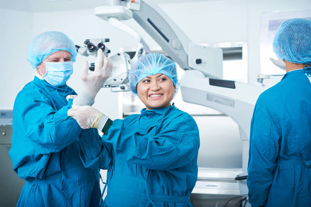 Aziatische chirurgisch assistent glimlachen en op zoek weg terwijl het helpen van de chirurg op latex handschoenen - Foto, afbeelding