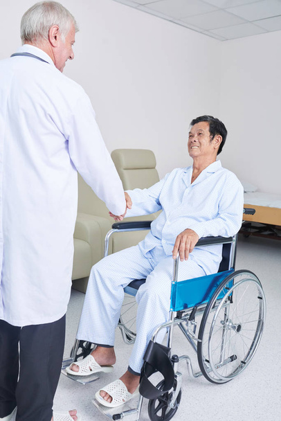 Ηλικιωμένοι Ασιατική άνθρωπο σε αναπηρικό αμαξίδιο, χαμογελώντας και κουνώντας το χέρι του ανώτερος ιατρού σε θάλαμο της σύγχρονης κλινικής - Φωτογραφία, εικόνα