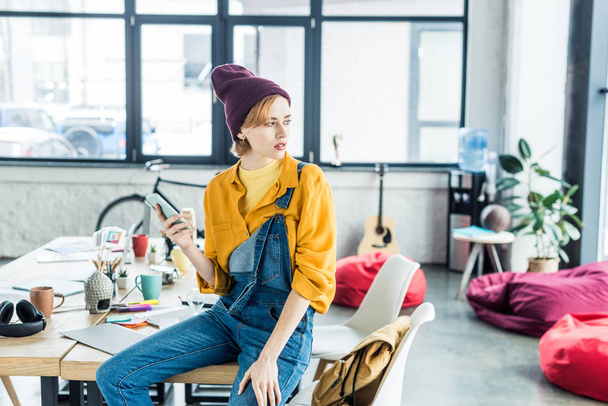 female designer sitting on table and using smartphone in loft office with copy space - Фото, изображение