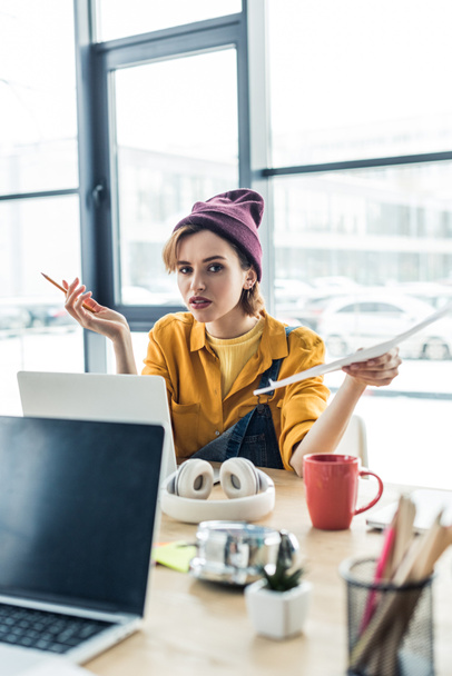 confused young female it specialist holding document and pen while sitting at computer desk in loft office  - Photo, Image