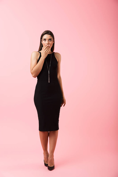 Full length portrait of a shocked beautiful young woman wearing black dress standing isolated over pink background - Fotografie, Obrázek