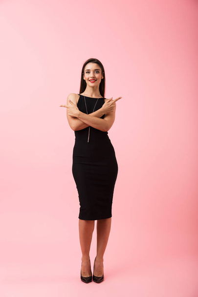Full length portrait of a beautiful young woman wearing black dress standing isolated over pink background, pointing at copy space - 写真・画像