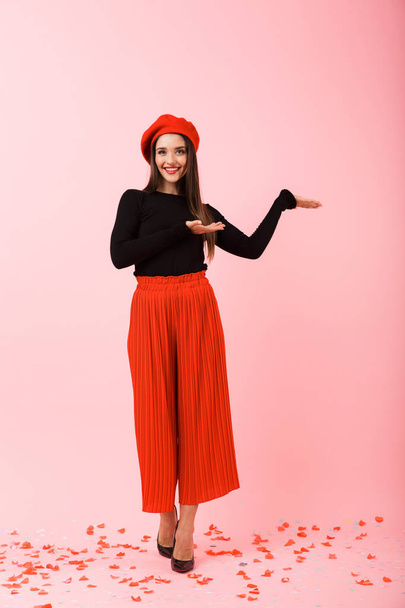Full length portrait of a beautiful young woman wearing red beret standing isolated over pink background, presenting copy space - Foto, Imagen
