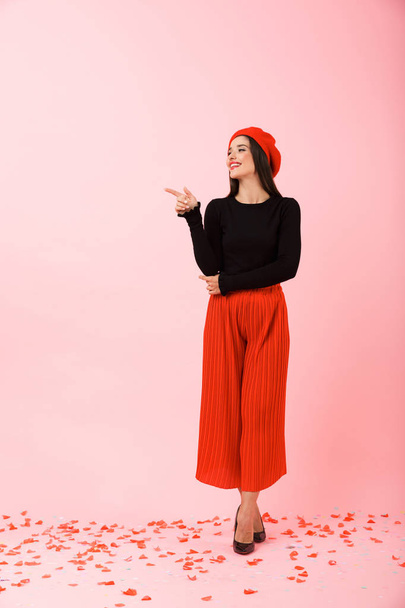 Full length portrait of a beautiful young woman wearing red beret standing isolated over pink background, presenting copy space - Foto, Bild